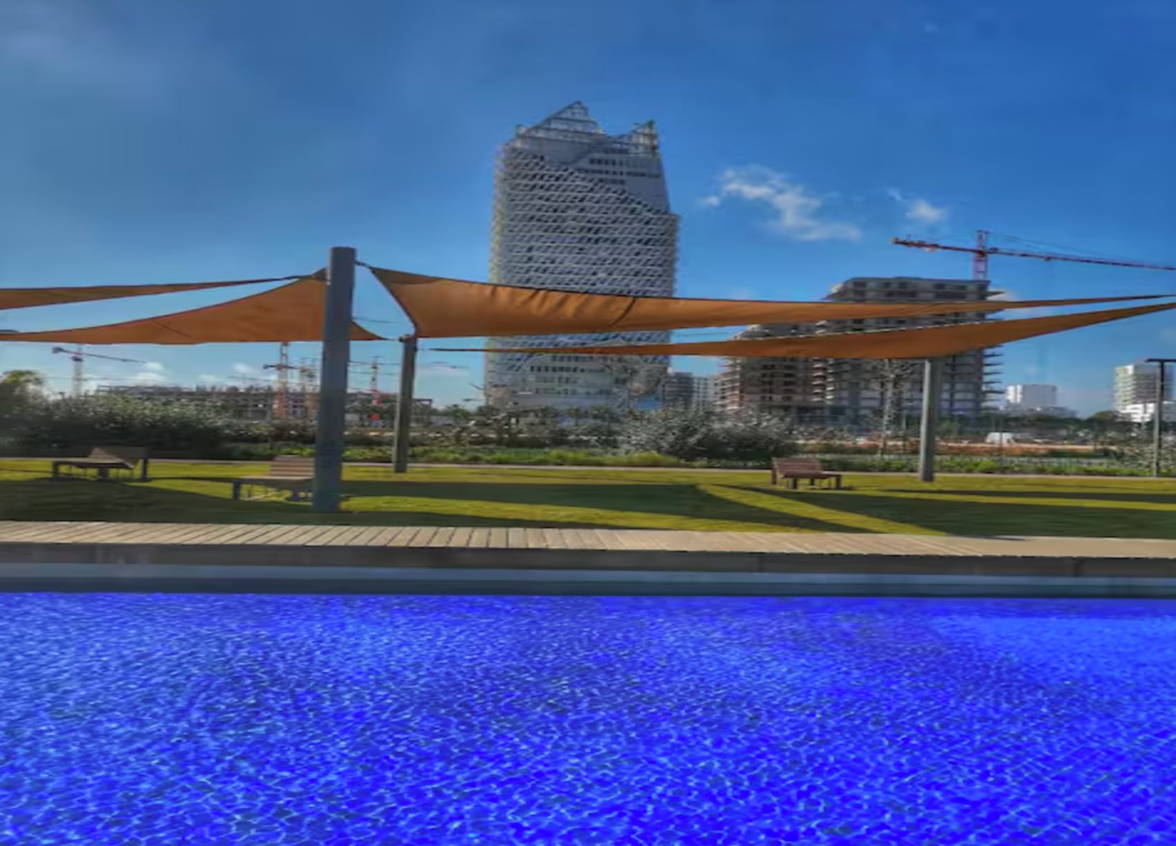 A swimming pool with a blue water surface, surrounded by a park with benches and a tall building in the background.