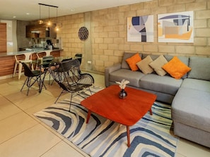Un salon moderne avec un canapé, une table basse et des fauteuils. On aperçoit un coin cuisine au fond.