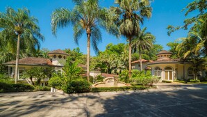 A residential area with a paved driveway leading to a gated entrance, surrounded by palm trees and a well-maintained lawn. - Harmon House 6 BR with private pool (Playa Herradura)
