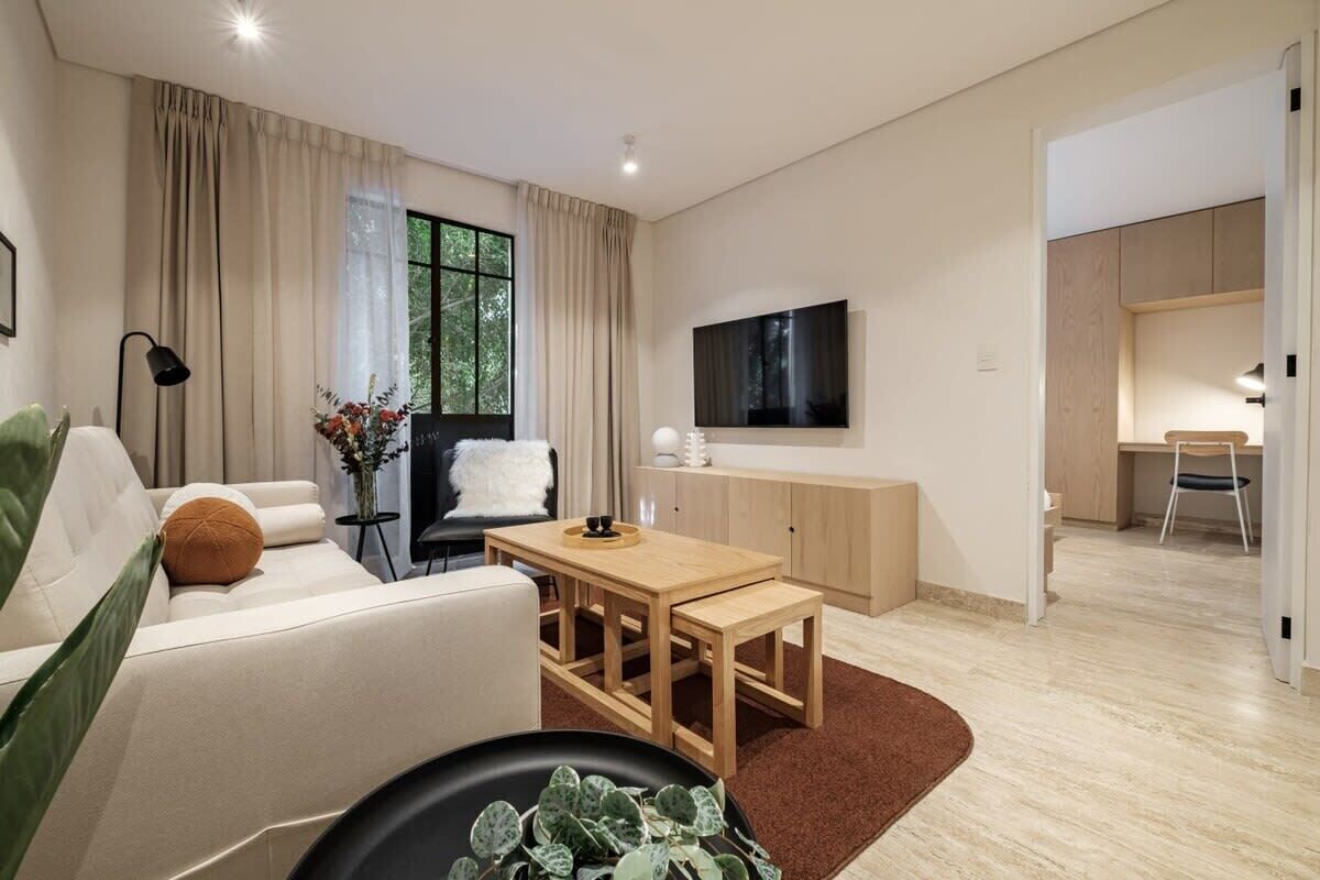 A modern living room with a sofa, a coffee table, a TV mounted on the wall, and a dining area with a table and chairs.