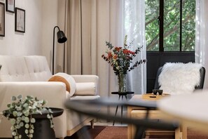 A modern living room with a sofa, a coffee table, a lamp, a vase with flowers, and a view of greenery through the window. - CAM 1-Bedroom Deluxe (Ciudad de México)