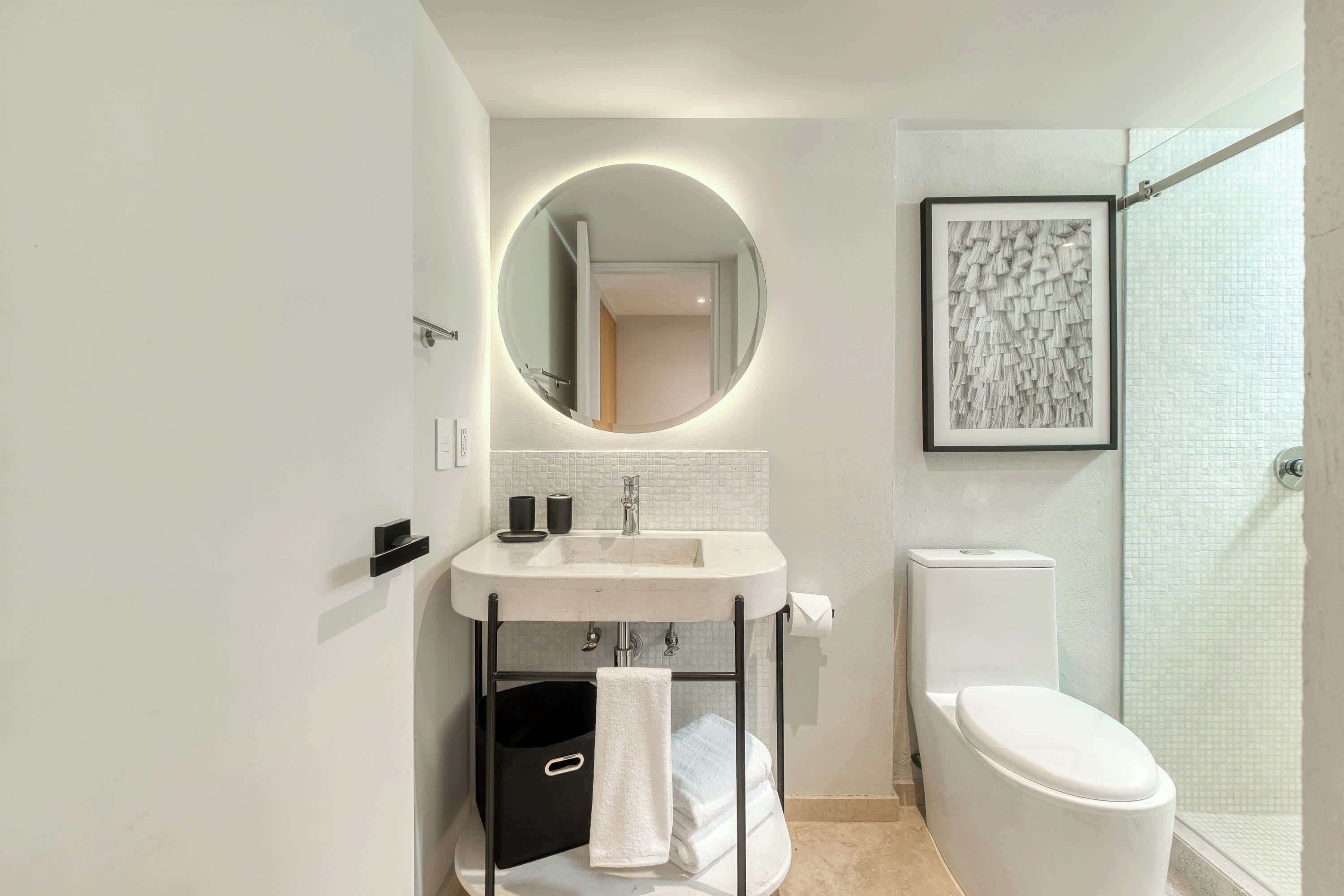 Ein modernes Badezimmer mit rundem Spiegel, weißem Waschbecken, Toilette und einer Duschkabine aus Glas.