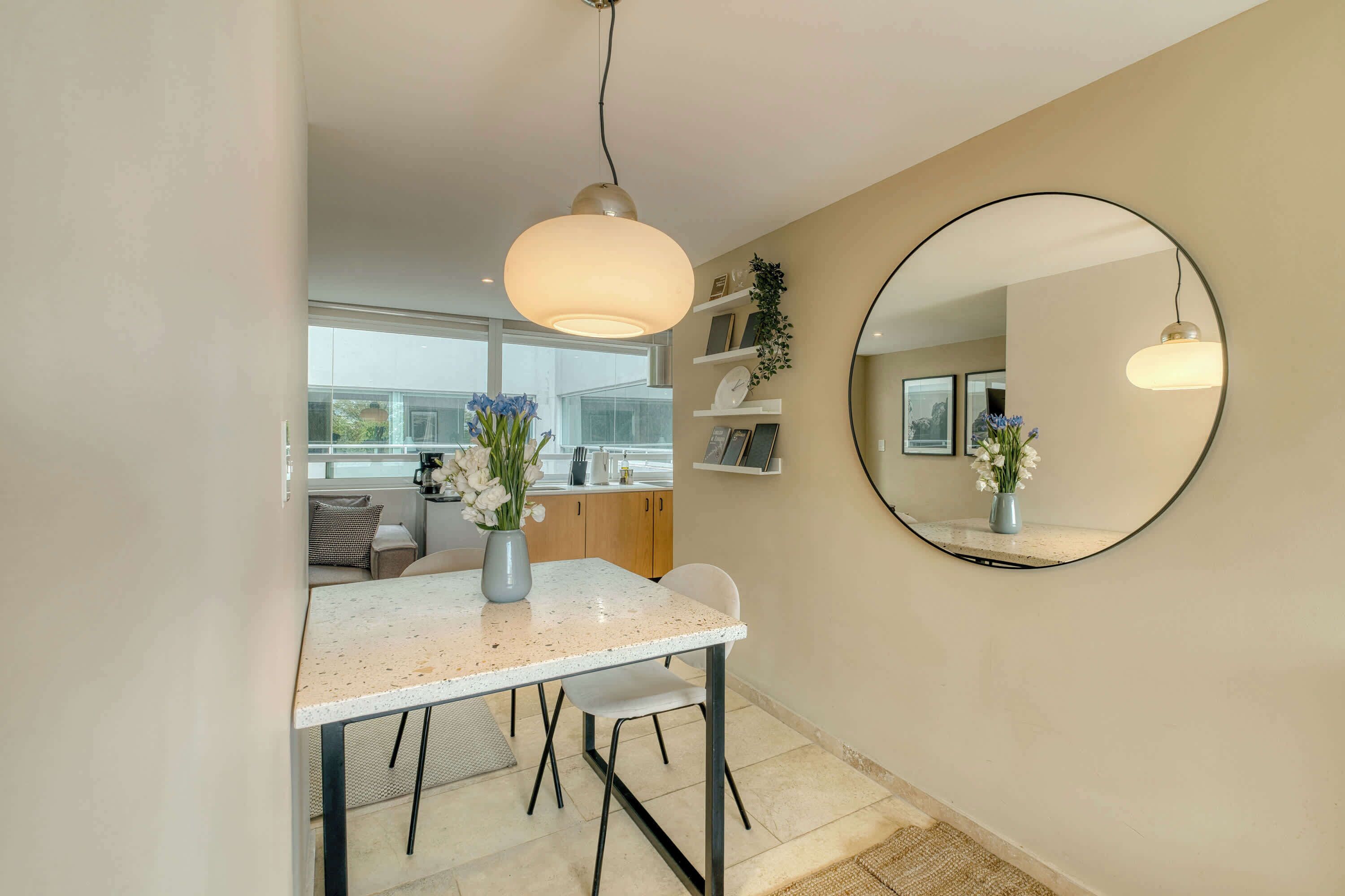 A modern kitchen with a dining area, a round mirror, and a vase with flowers.