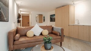 Una sala moderna con un sofá de cuero marrón, una mesa de centro de vidrio con una planta, y una cocina con armarios de madera y encimera blanca.