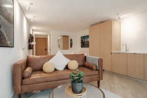 Ein modernes Wohnzimmer mit einem braunen Ledersofa, einem Glastisch mit einer Pflanze und einer Küchenzeile mit Holdekor-Schränken und einer weißen Arbeitsplatte.