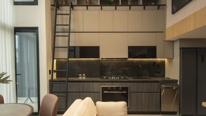 Cocina moderna con estufa, horno y lavavajillas integrados. Cuenta con una zona de comedor con mesa y sillas, además de un sofá. Se ve una escalera en la cocina y una planta sobre la encimera.