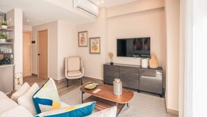 Ein modernes Wohnzimmer mit einer Couch, einem Couchtisch und einem Fernseher. Eine Tür führt in einen weiteren Raum, und ein kleines Sideboard beherbergt dekorative Gegenstände.