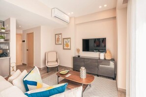 Ein modernes Wohnzimmer mit einer Couch, einem Couchtisch und einem Fernseher. Eine Tür führt in einen weiteren Raum, und ein kleines Sideboard beherbergt dekorative Gegenstände.