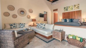 Ein geräumiges Zimmer mit einer Couch, einem Korbsessel, einem Nachttisch, einem Fernseher und einem Bett mit Holz Kopfteil.