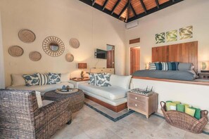 Ein geräumiges Zimmer mit einer Couch, einem Korbsessel, einem Nachttisch, einem Fernseher und einem Bett mit Holz Kopfteil.