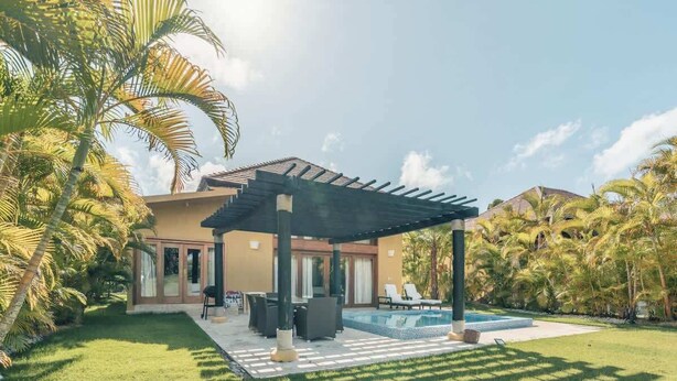 Ein modernes Haus mit überdachter Terrasse, Swimmingpool und üppiger Bepflanzung.