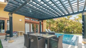 Eine Terrasse im Freien mit Tisch aus Glasplatten, Korbstühlen, einem Pool und einem überdachten Bereich mit einer Pergola.