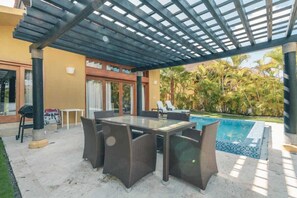 Eine Terrasse im Freien mit Tisch aus Glasplatten, Korbstühlen, einem Pool und einem überdachten Bereich mit einer Pergola.