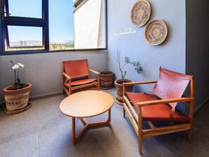 Ein Zimmer mit großem Fenster, einem runden Holztisch, zwei Holzsesseln mit orangefarbenen Kissen und einem Blumentopf.