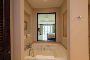 Een badkamer met een bad, een douche en een spiegel die een slaapkamer met een bed en gordijnen weerspiegelt.
