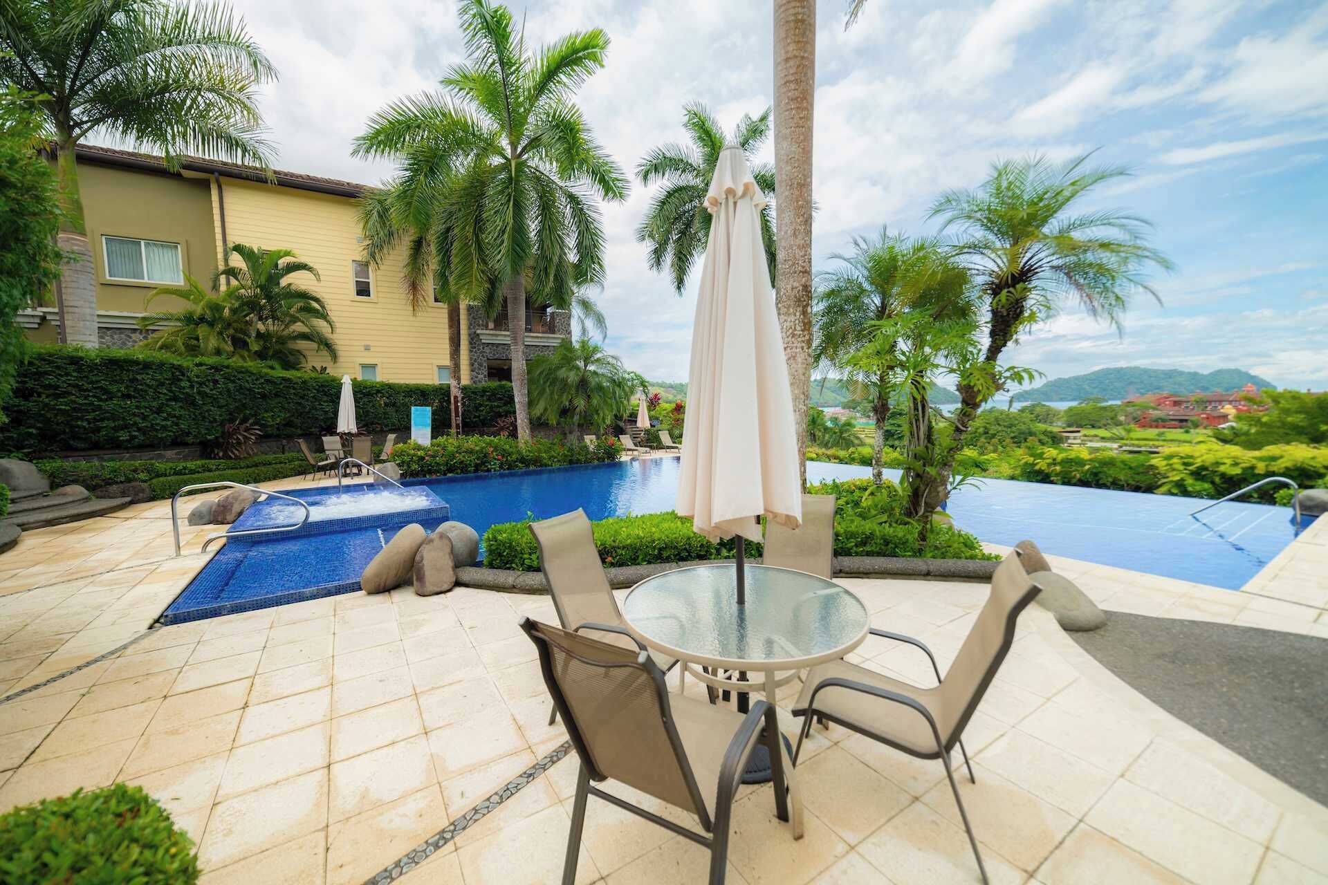 A poolside area with a glass table and chairs, a hot tub, and a view of a mountain and a building in the distance.