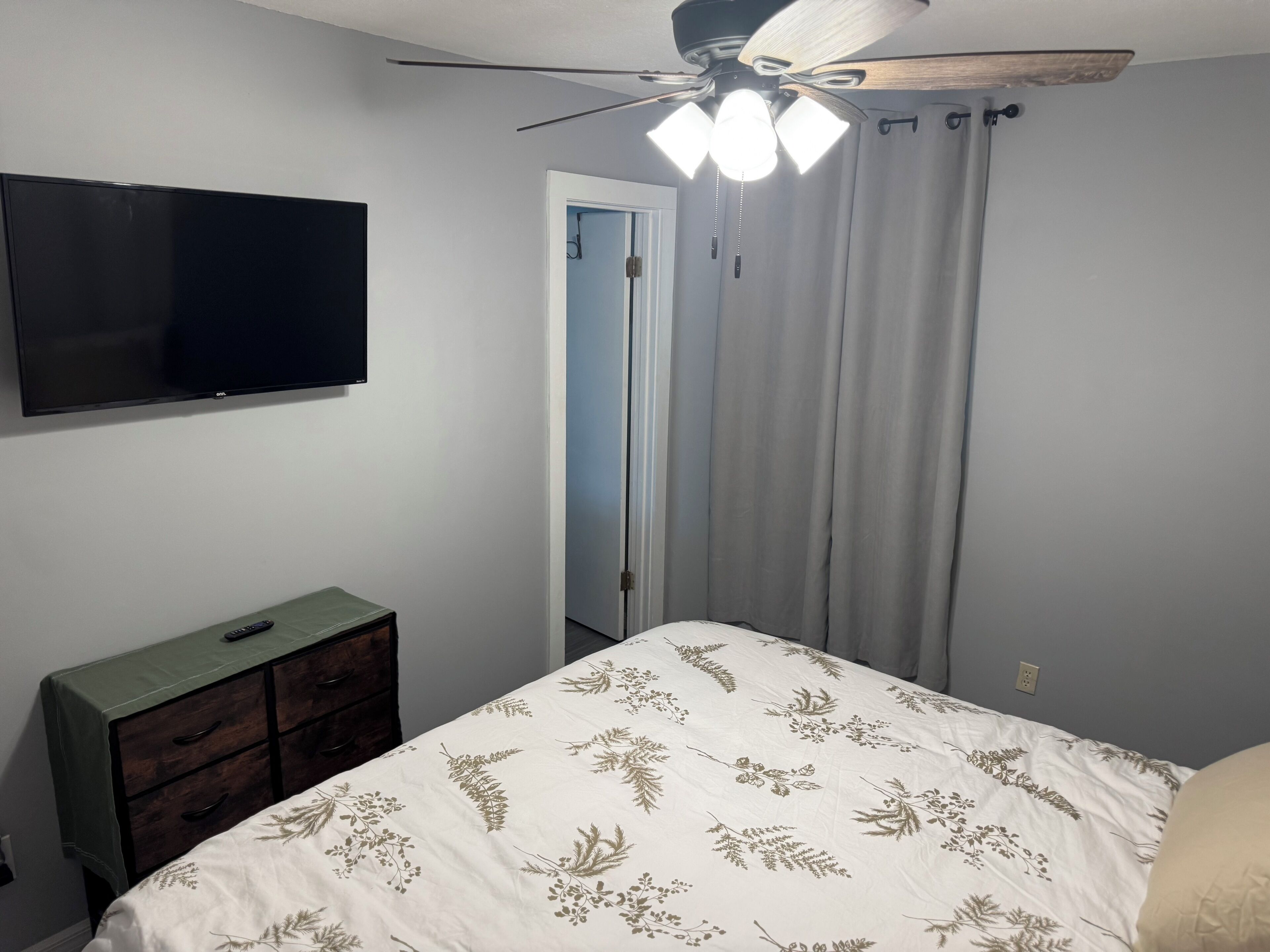 A bedroom with a bed, a dresser, a ceiling fan, a television, and a door.