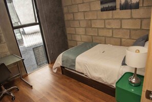 Ein Schlafzimmer mit Bett, Schreibtisch, Stuhl, Lampe und Blick durch ein Fenster.