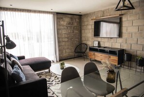 Ein modernes Wohnzimmer mit einer Couch, einem Couchtisch, einem an einer Ziegelwand montierten Fernseher und einem Essbereich mit Stühlen.