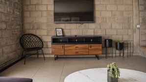 Ein Wohnzimmer mit an einer Ziegelwand montiertem Fernseher, einem Holzschrank, einem schwarzen Sessel und einem runden Tisch mit einer Pflanze.
