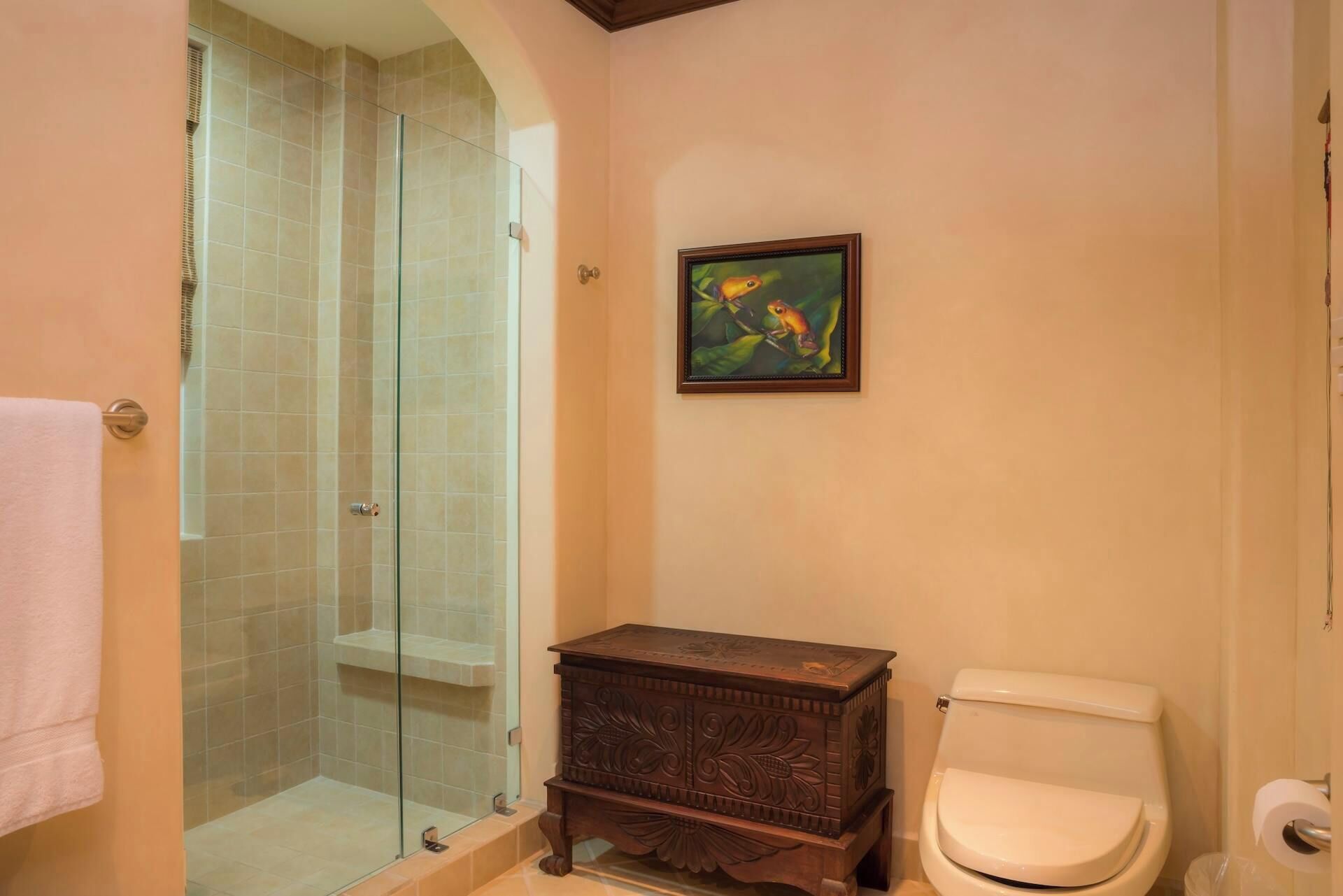 Une salle de bain avec une douche à parois de verre, une commode en bois, des toilettes et un tableau encadré au mur.