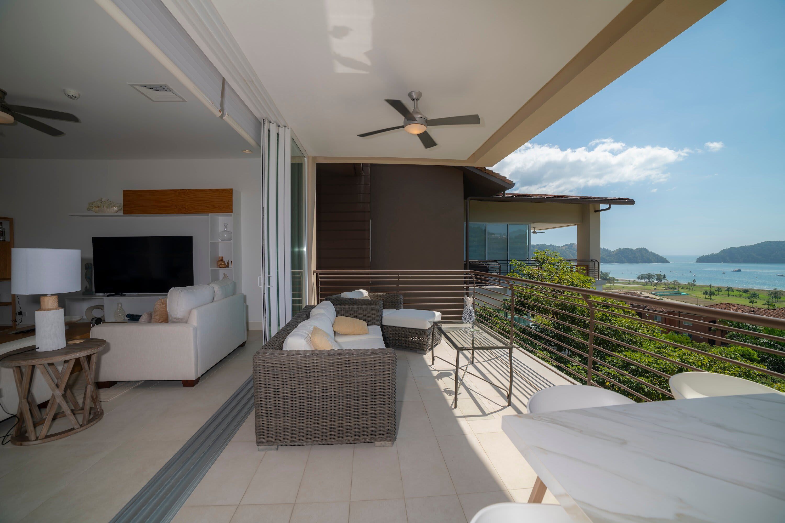 A balcony with a TV, sofa, and dining area overlooking a scenic view.