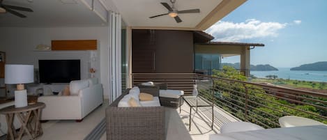 A balcony with a TV, sofa, and dining area overlooking a scenic view.