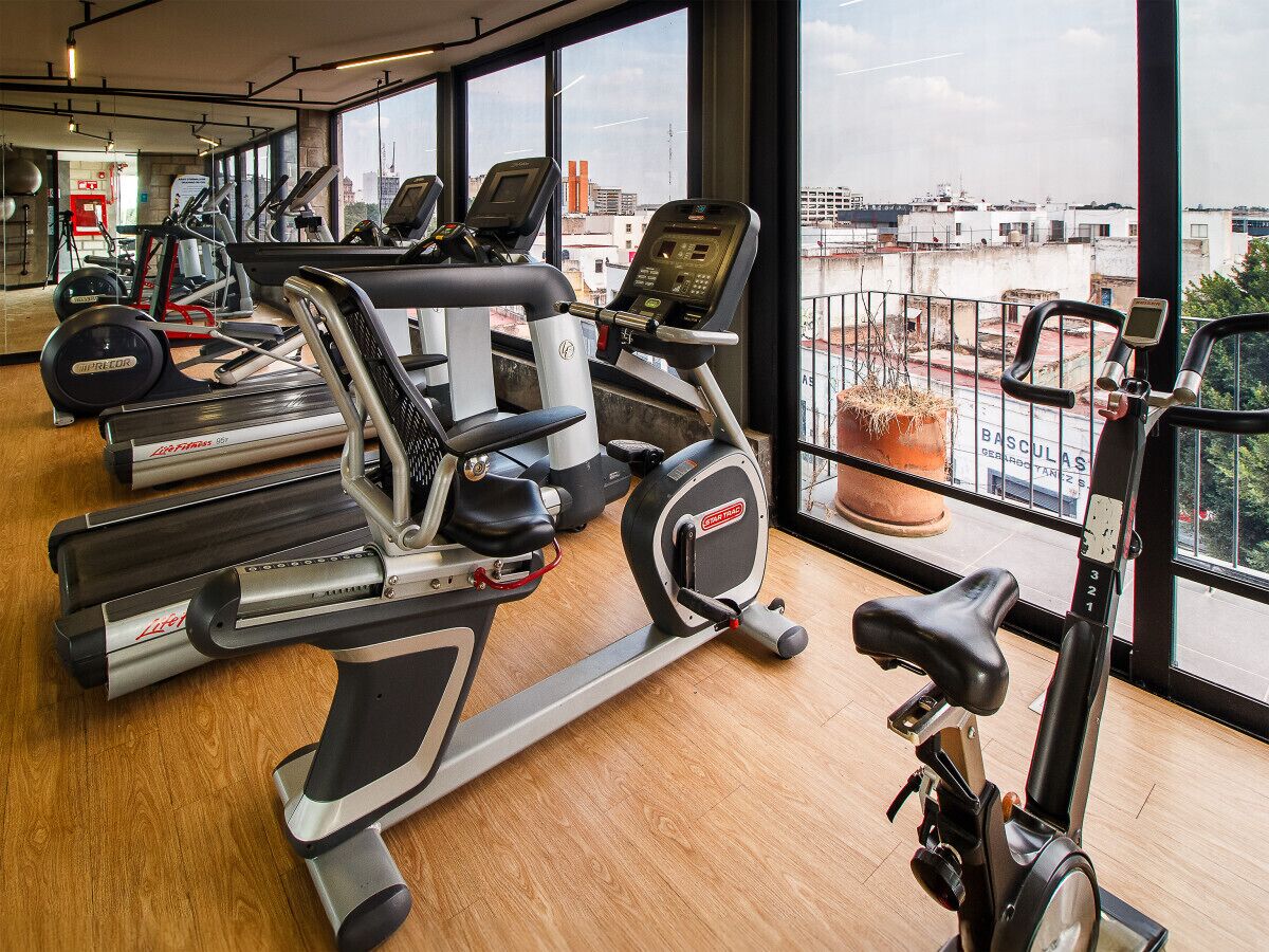 A gym with cardio machines, a weight bench, and a view of the city through large windows.