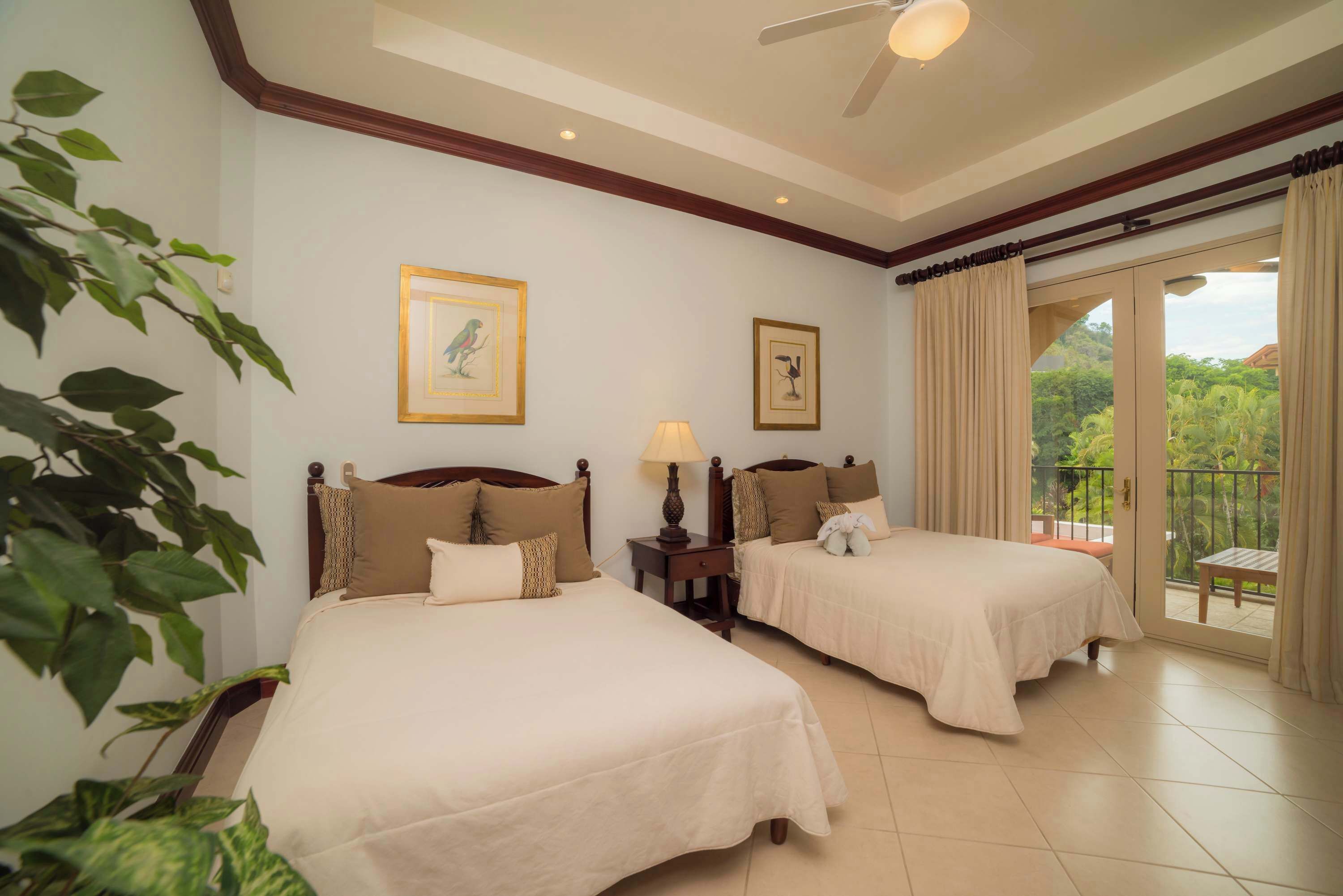 A hotel room with two beds, a ceiling fan, a balcony with curtains, and framed artwork on the walls.