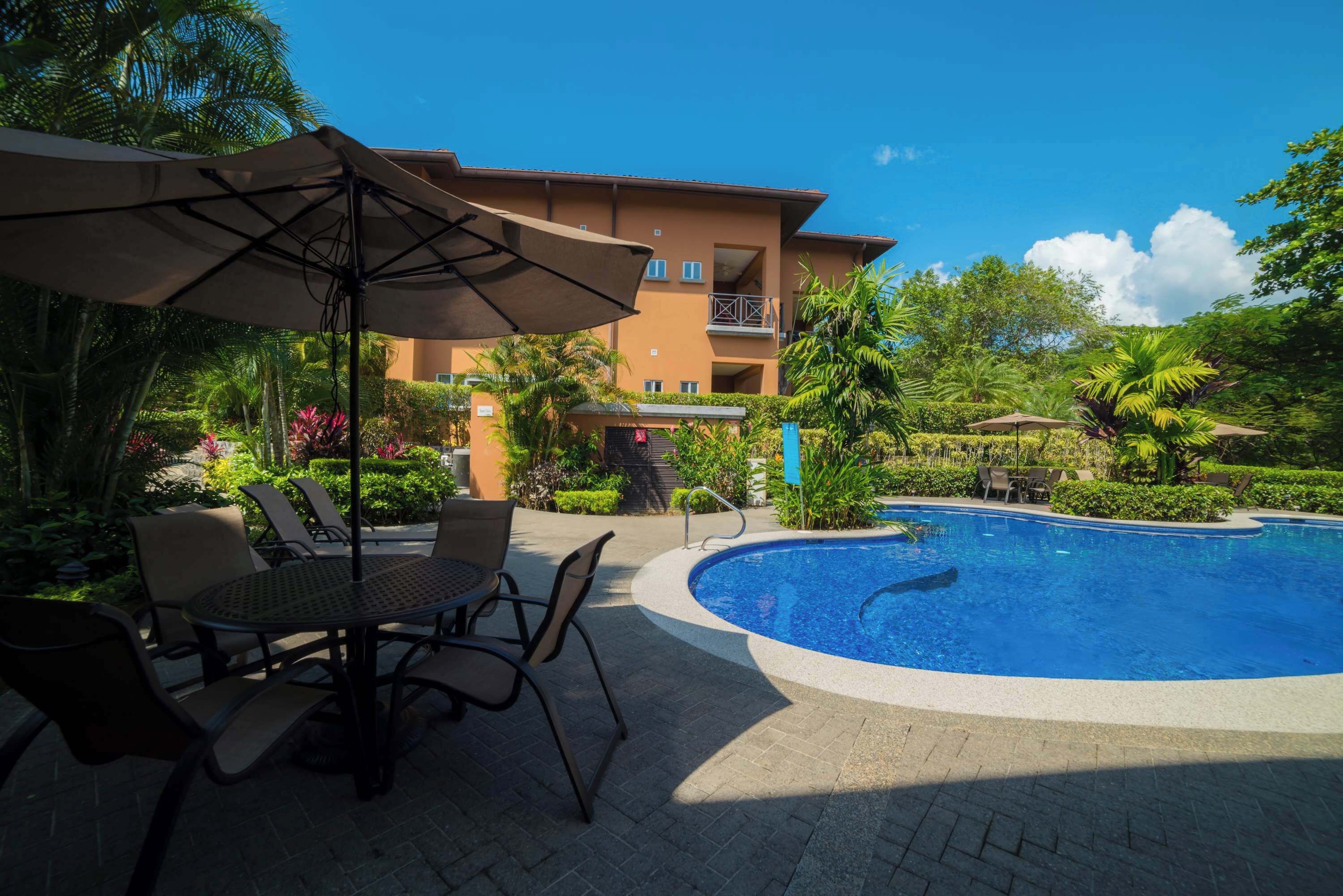 Zona de piscina con sillones de descanso, una mesa y una sombrilla grande.