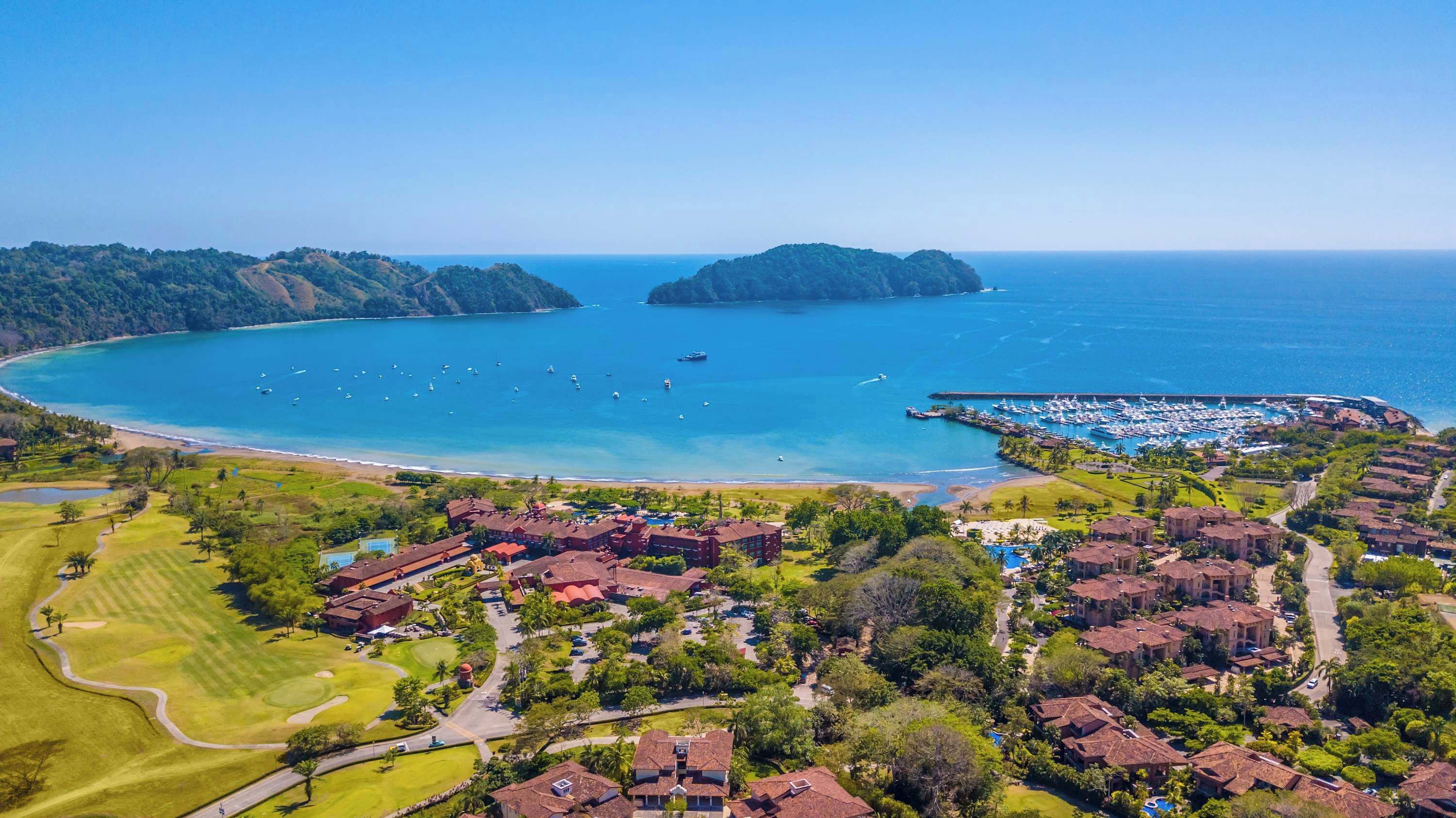 Vista aérea de un complejo turístico costero con un puerto deportivo, campo de golf y exuberante vegetación.