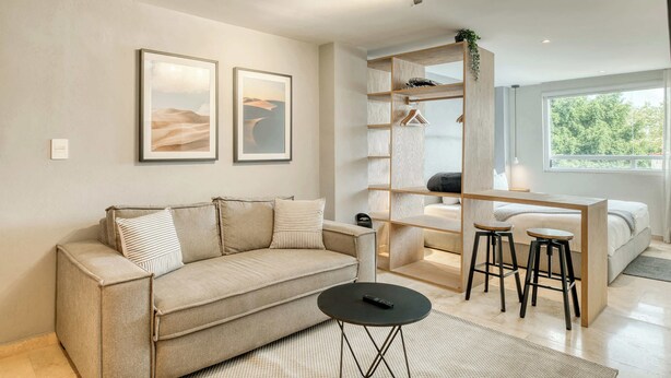 A modern living room with a sofa, a coffee table, a desk with two stools, and a bed. There are two framed pictures on the wall, a plant on the desk, and a large window providing natural light. - Modern King Studio in Central Roma Norte (Ciudad de México)
