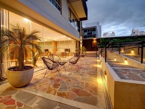 Ein modernes Hotel mit einer Steinterrasse, Sitzgelegenheiten im Freien und einer großen Kübelpalme.