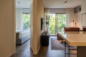 Ein modernes Hotelzimmer mit Bett, Essbereich, kleinem Tisch mit Pflanze und Blick auf Grünfläche.