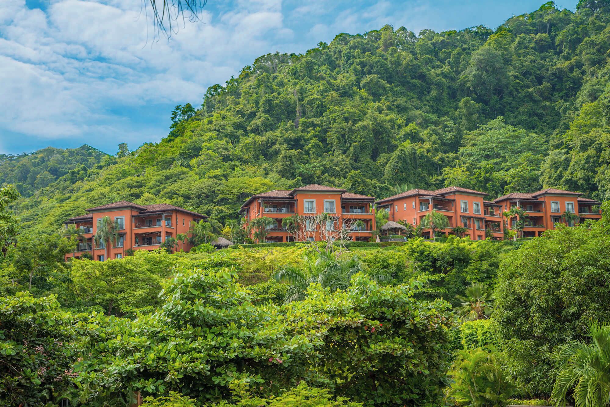 A row of red buildings with balconies, surrounded by lush greenery and a forested hillside.
