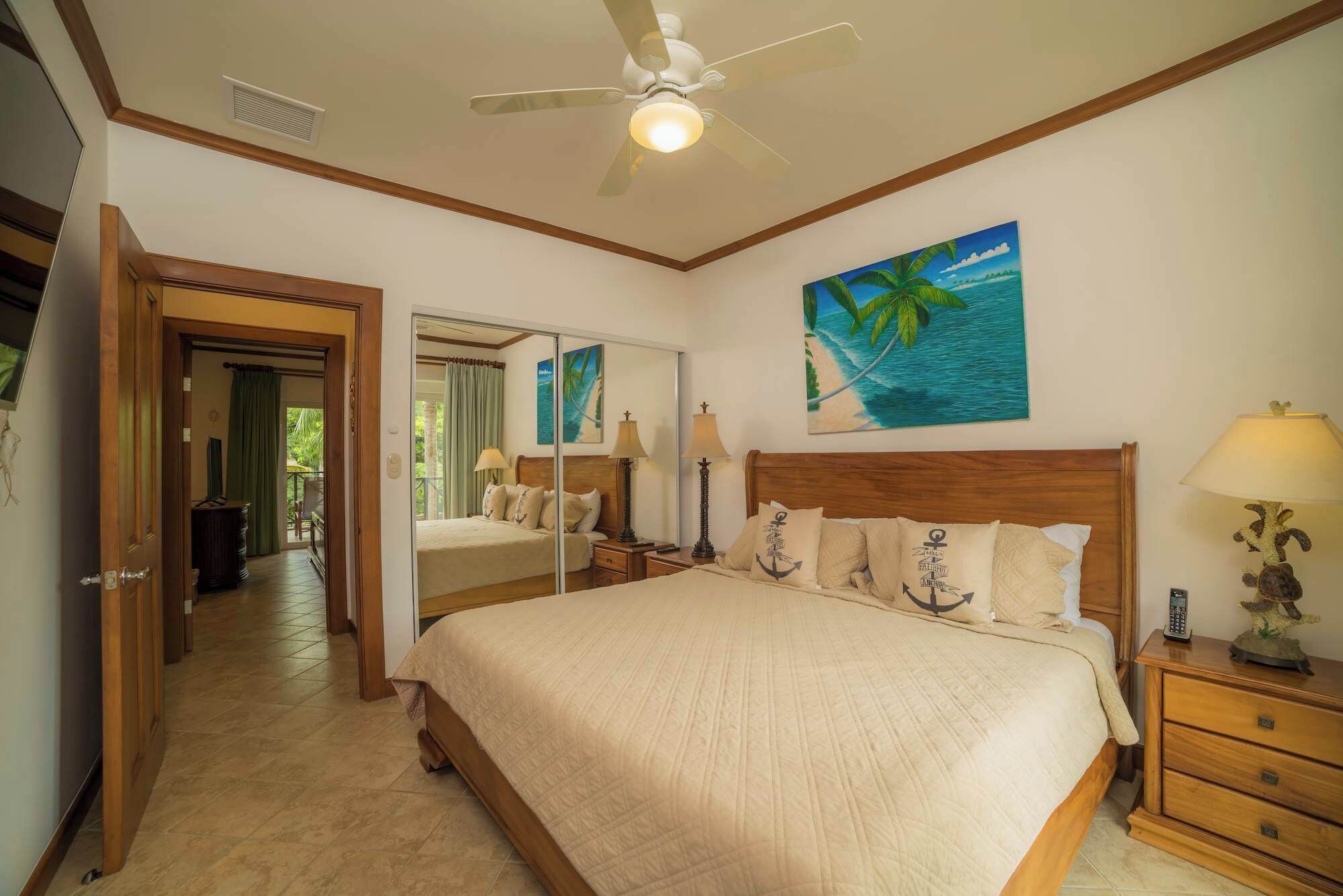 A bedroom with a bed, bedside tables, a ceiling fan, a painting, and a view of a balcony.