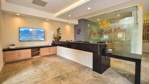Ein Empfangsbereich mit einem schwarzen Empfangstresen, einem Wand-Fernseher und einer Glaswand mit dem Schriftzug 'Stay in Costa Rica'.