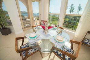 A dining area with a glass table and chairs, a view of the sea, and a balcony with a railing.