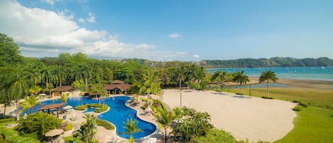Un resort con una amplia piscina, una playa de arena y abundante vegetación.
