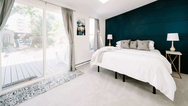 A bedroom with a bed, a nightstand, a lamp, a window with curtains, and a view of the outdoors. - Olde Town Ambler A Distinctive Walkable Retreat (Arvada)
