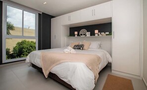 Ein Schlafzimmer mit großem Fenster, einem Bett mit weißer Bettwäsche, einem Regal mit Dekoration und einer dunklen Wand.