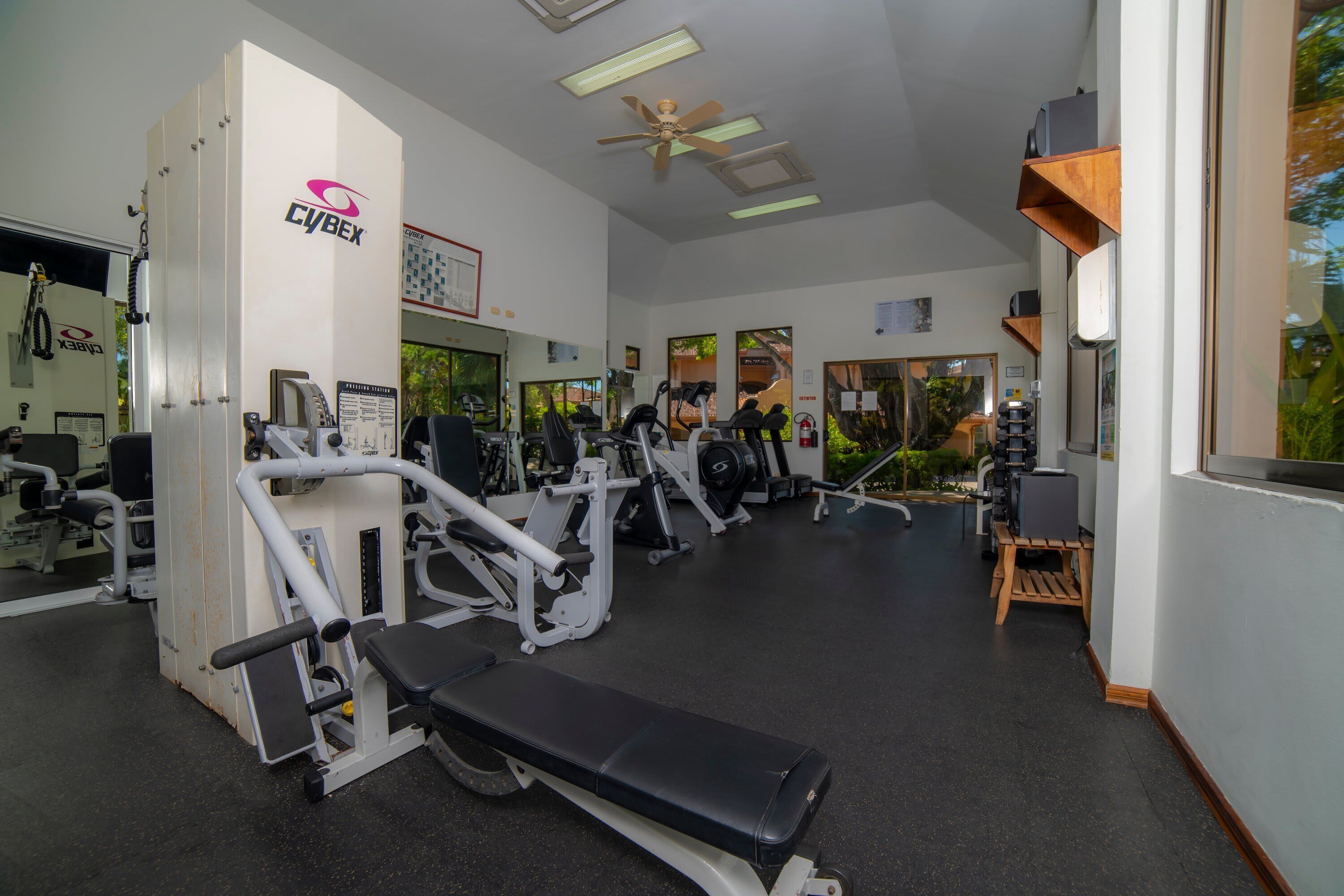 A gym with various exercise machines, a ceiling fan, and large windows.