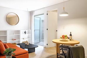 Ein modernes Wohnzimmer mit einer Couch, einem Couchtisch, einem runden Tisch mit einer Obstschale und einer Flasche sowie einem großen Spiegel an der Wand.