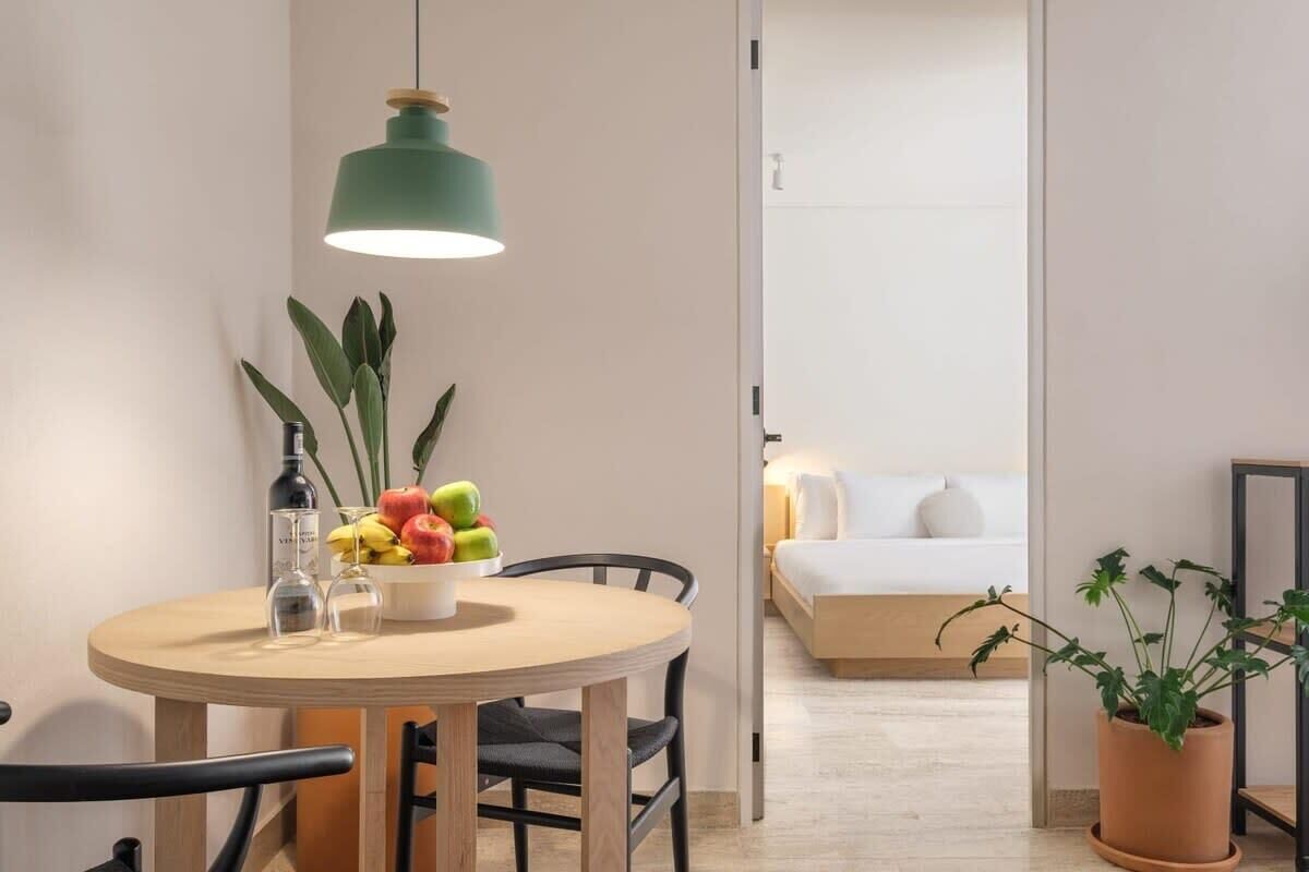 A compact room with a round dining table, chairs, a hanging light fixture, a potted plant, and a bed with a white headboard.