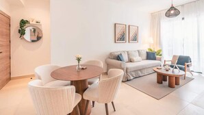 A modern living room with a round wooden coffee table, a sofa, and two armchairs. There is a dining table with chairs, a mirror, and framed artwork on the wall. - Cozy 1br with Balcony, Pool & Beach Access Cap Cana (Punta Cana)