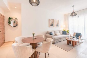 Ein modernes Wohnzimmer mit einem runden Holztisch, einer Couch und zwei Sesseln. Es gibt einen Esstisch mit Stühlen, einen Spiegel und gerahmte Kunstwerke an der Wand.