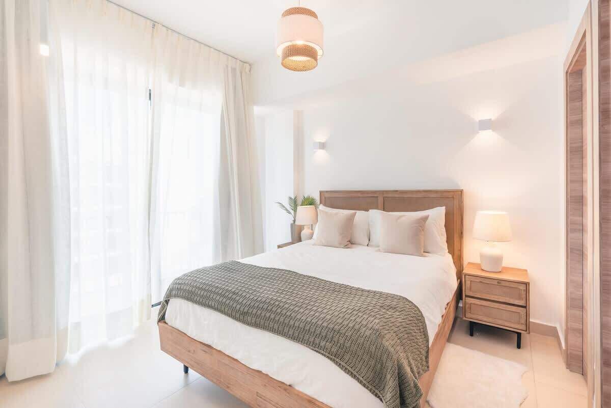 A bedroom with a wooden bed, white bedding, a bedside table with a lamp, and a large window with sheer curtains.