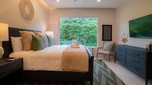 Quarto com cama, mesa de cabeceira, penteadeira, cadeira e uma janela grande com vista para árvores.