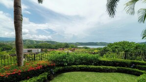 A landscaped garden with a curved pathway, a palm tree, and a view of a coastal area with buildings in the distance. - Montebello 1E 3 bed 3 bath, ocean view (Playa Herradura)