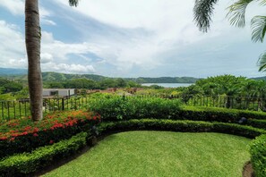 A landscaped garden with a curved pathway, a palm tree, and a view of a coastal area with buildings in the distance. - Montebello 1E 3 bed 3 bath, ocean view (Playa Herradura)